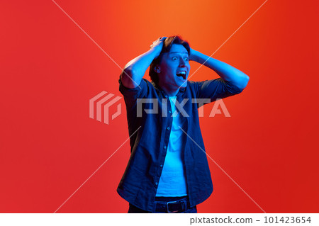 Studio shot of young guy, student screaming with exciting face over red-orange background in neon light. Amazement 101423654