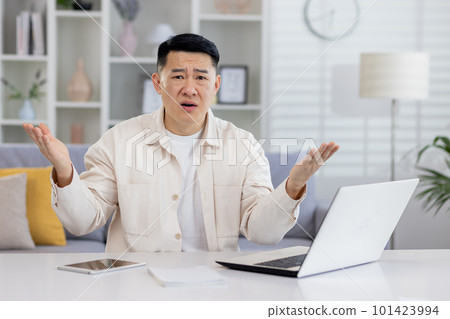 Worried Asian man working at home on laptop, looking confused at camera, spreading hands. Financial problems, broken device, remote work. Worried Asian man working at home on laptop, looking confused at camera, spreading hands. Financial problems, broken device, remote work. 101423994