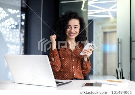 A young Latin American business woman is sitting in the office and holding the phone in her hands. A successful financial deal rejoices, celebrates, shows victory with his hand, looks at the camera. 101424005