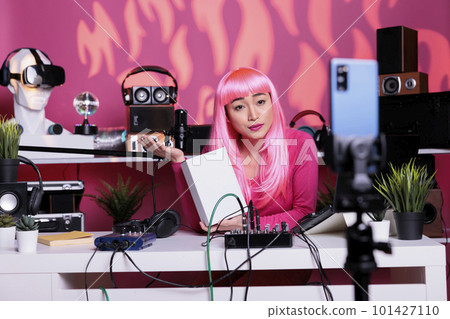 Asian vlogger with pink hair holding white box advertising product in front of smartphone camera, filming review for vlogging channel. Influencer recording vlog using professional equipment 101427110