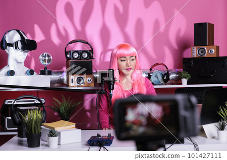 Smiling asian vlogger with pink hair talking with subscribers while recording vlog using professional camera, posting podcast on social media. Positive influencer filming video content 101427111