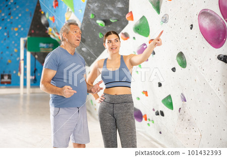 Friendly young female coach of climbing gym instructing aged man 101432393