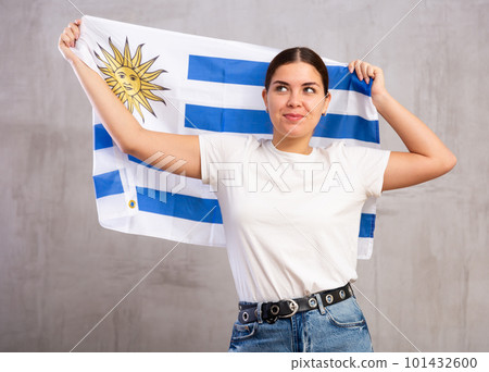 Excited young woman in casual wear waving state flag of Uruguay against gray wall background indoors Excited young woman in casual wear waving state flag of Uruguay against gray wall background indoors 101432600
