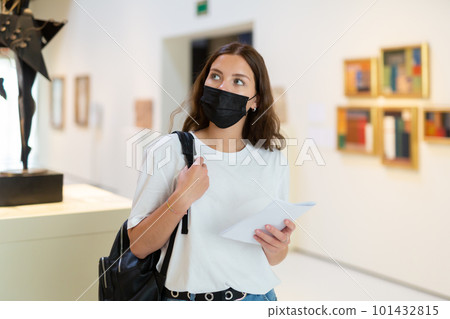 Young girl wearing a protective mask in the museum looks at the exhibits 101432815