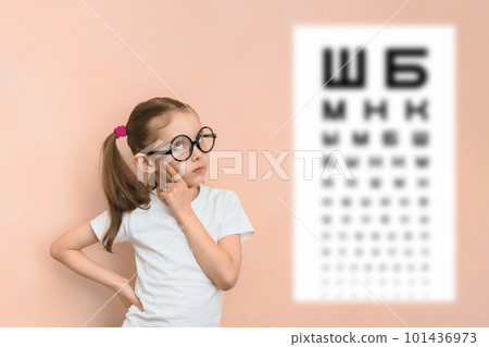 Thoughtful smart girl of elementary school age in round glasses against the background of a blurred sivtsev table for checking visual acuity 101436973