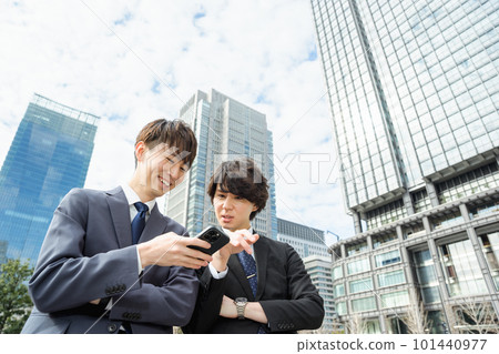 Two men in their 20s and 30s in suits looking at their smartphones and having a conversation with a smile in a city surrounded by skyscrapers Two men in their 20s and 30s in suits looking at their smartphones and having a conversation with a smile in a city surrounded by skyscrapers 101440977
