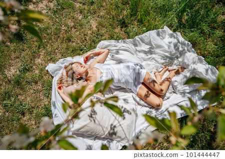 woman sleeps on a white bed in the fresh spring grass in the garden. Dressed in a blue nightgown. woman sleeps on a white bed in the fresh spring grass in the garden. Dressed in a blue nightgown. 101444447