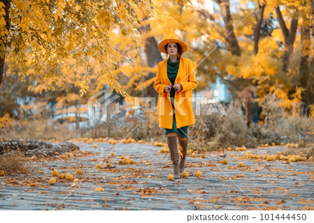 Beautiful woman walks outdoors in autumn. She is wearing a yellow coat, yellow hat and green dress. Young woman enjoying the autumn weather. Autumn content Beautiful woman walks outdoors in autumn. She is wearing a yellow coat, yellow hat and green dress. Young woman enjoying the autumn weather. Autumn content 101444450