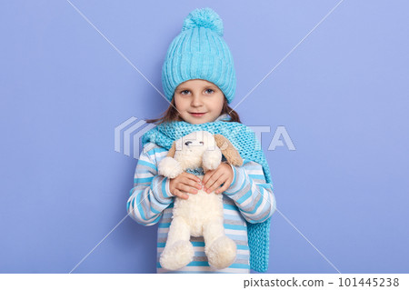 Image of charming little female, child with fluffy puppy in hands, looking directly at camera, dresses casual clothing, scarf and hat, posing isolated over lilac studio background. Childhood concept. 101445238
