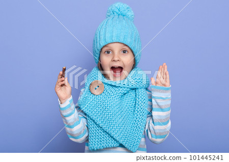 Beautiful little girl expressing joyous emotions, keeps ahnds up and mouth opened, child wearing casual striped shirt, scarf and cap with pom pom, holding candy in hand, isolated over blue background. Beautiful little girl expressing joyous emotions, keeps ahnds up and mouth opened, child wearing casual striped shirt, scarf and cap with pom pom, holding candy in hand, isolated over blue background. 101445241