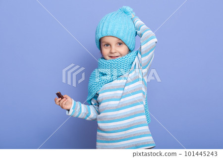 Charming astionish little girl looking at camera with surprised facial expression isolated over blue studio background, child holding candy in hand and keeps hand on head, wears cap, scarf and shirt. Charming astionish little girl looking at camera with surprised facial expression isolated over blue studio background, child holding candy in hand and keeps hand on head, wears cap, scarf and shirt. 101445243