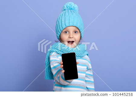 Astonished little girl kid wearing winter hat with pom pom showing blank screen for text space of new popular mobile phone isolated over blue background, child standing with opened mouth and surprised 101445258