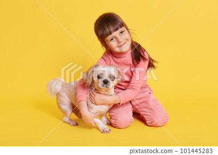 Cute dark haired girl playing with pet dog, looking directly at camera with smile, isolated over yellow background, charming female child with puppy, wearing rose casual attire. Cute dark haired girl playing with pet dog, looking directly at camera with smile, isolated over yellow background, charming female child with puppy, wearing rose casual attire. 101445283