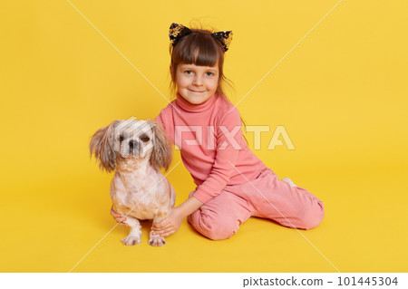 Little dark haired girl with puppy posing indoor, child looking at camera and smiling, hugging her pet, wears casual attire and hair band with cat's ears, isolated over yellow background. 101445304
