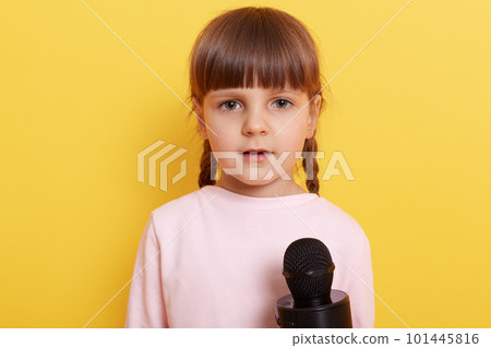 Cute shy child wearing pale pink shirt speaking in microphone, looks at camera with a bit confused look, small kid with pigtails being interviewed against yellow wall. 101445816