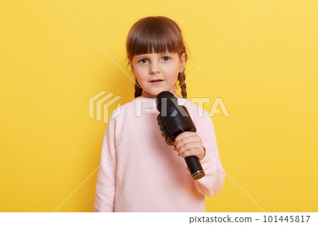 Adorable little girl with microphone on yellow background, looks at camera while talking in mic, pointing index finger aside. Copy pace for advertisement or promotional text. 101445817