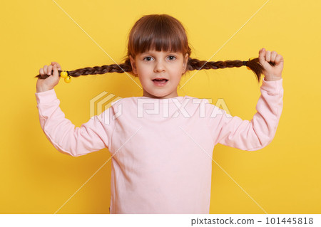Cute girl pull her pigtails aside by hands, looks at camera with opened mouth, Caucasian female kid in pale pink shirt against yellow wall, child having alone. 101445818