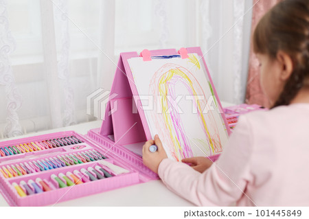 Talented child girl draws rainbow with colored pencils while sitting at table with set of art and creativity, has lots colors of wax crayons, making illustration. 101445849