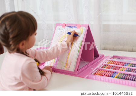 Little girl with dark hair drawing with colourful pencils on canvas, has great variety of multicolored pencils and wax crayons, kid sitting at table in front of window. 101445851
