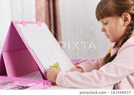 Talented child girl draws with colored pencils, being concentrated and diligent, sitting at desk with set of art and creativity, kid wearing casual attire, having pigtails painting at home. 101446637