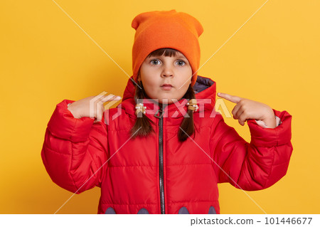 Small girl looking at camera with big eyes, pointing at her cheeks with her index fingers, making funny grimace with her lips isolated over yellow background, kid wearing red jacket and orange cap. Small girl looking at camera with big eyes, pointing at her cheeks with her index fingers, making funny grimace with her lips isolated over yellow background, kid wearing red jacket and orange cap. 101446677