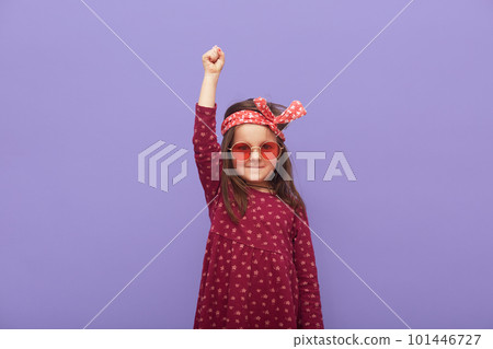 Little fashionable rebel girl dressed in dress, hair band and stylish glasses looks at camera with a grave expression on her face and raised fist up, child defends her rights on purple background Little fashionable rebel girl dressed in dress, hair band and stylish glasses looks at camera with a grave expression on her face and raised fist up, child defends her rights on purple background 101446727