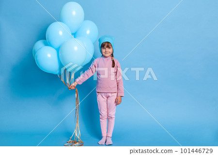 Dark haired little girl in cap and pink suit, holds big bunch of helium balloons, being on children's party, looks shyly at camera, full length of female child celebrates birthday. 101446729