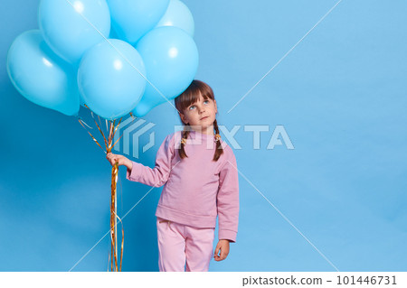 Cute European female child wearing rose closing, kid with pigtails looking way with thoughtful facial expression, dreams about something pleasant, holding bunch of helium balloons, against blue wall. 101446731