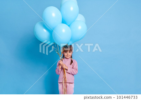 Charming small girl looks attentively at camera, holding bunch of helium balloons, wears pink jumper and trousers, prepares to celebrate birthday, models over blue background, goes for kid's party. Charming small girl looks attentively at camera, holding bunch of helium balloons, wears pink jumper and trousers, prepares to celebrate birthday, models over blue background, goes for kid's party. 101446733