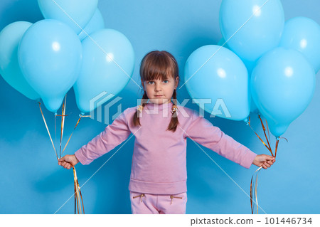 Caucasian attractive female kid goes on birthday party, carries blue helium balloons in both hands, wears casual rose jumper, isolated on color background. Caucasian attractive female kid goes on birthday party, carries blue helium balloons in both hands, wears casual rose jumper, isolated on color background. 101446734