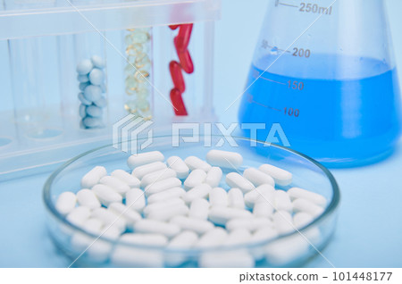 Selective focus on pile of white medical pills in petri dish against labware with capsules and blue solution, isolated on blue background. World Health Day. Pharmaceutical industry. Clinical research 101448177