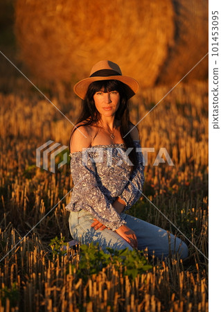 A woman with long black hair in jeans, a straw hat sits on her knees in a field 101453095