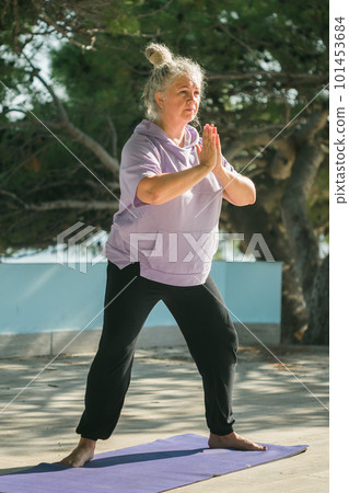 Serious aged woman in with dreadlocks hairstyle standing on beach against sea while practicing pose or asana during sunny day 101453684