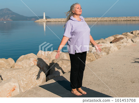 Serious aged woman in with dreadlocks hairstyle standing on beach against sea with raised arms while practicing pose during sunny day 101453708