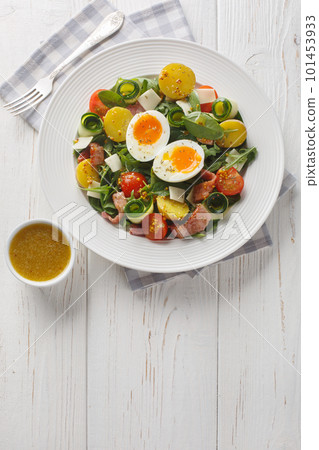 Fresh peasant salad with arugula, spinach, potatoes, bacon, tomatoes, cheese and soft-boiled eggs close-up on a plate. Vertical top view 101453933