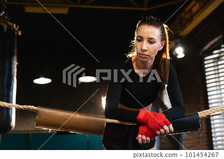 Woman fighter girl power. Woman fighter with red boxing wraps protective bandages standing on boxing ring leaning on ropes waiting resting. Strong powerful girl. Strength fit body workout training 101454267