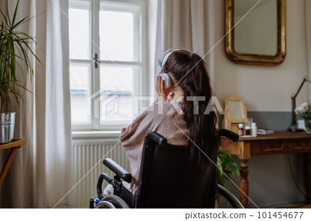 Teenage girl sitting on a wheelchair, listening music and looking out of the window. 101454677