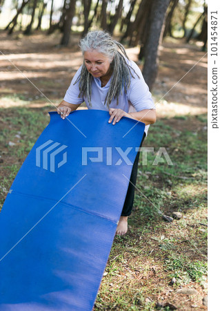 Old woman holding yoga mat and preparing to practice yoga or meditation outdoors on park. Happy mature woman with dreadlocks exercising. Meditation or yoga and relaxation concept Old woman holding yoga mat and preparing to practice yoga or meditation outdoors on park. Happy mature woman with dreadlocks exercising. Meditation or yoga and relaxation concept 101454871