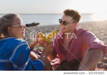 Happy young couple dating together in beach, sitting on a blanket and eating corn. Enjoying holiday time together. 101454897