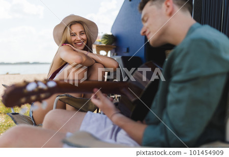 Young couple sitting together in front of van, camping and playing at guitair. 101454909