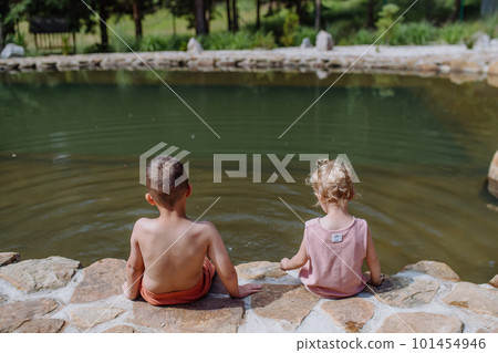 Cute girl and boy sitting together on the footpath by the lake, dangle their feet in the water, summer vacation concept. Cute girl and boy sitting together on the footpath by the lake, dangle their feet in the water, summer vacation concept. 101454946