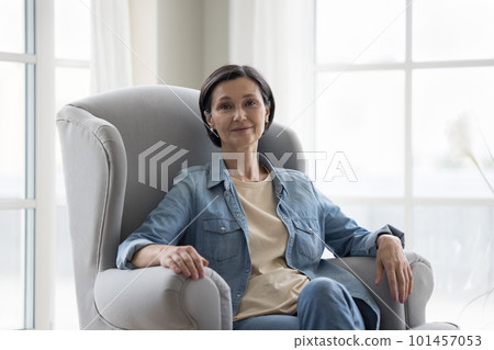 Positive senior 50s woman sitting in armchair, looking at camera 101457053