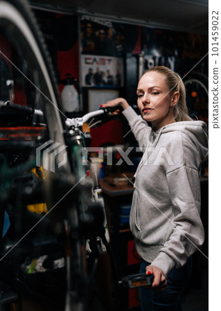 Vertical portrait of attractive bike mechanic female repairing and fixing mountain bicycle standing on bike rack in repair workshop with dark interior, looking at camera. Concept of bike maintenance. Vertical portrait of attractive bike mechanic female repairing and fixing mountain bicycle standing on bike rack in repair workshop with dark interior, looking at camera. Concept of bike maintenance. 101459022