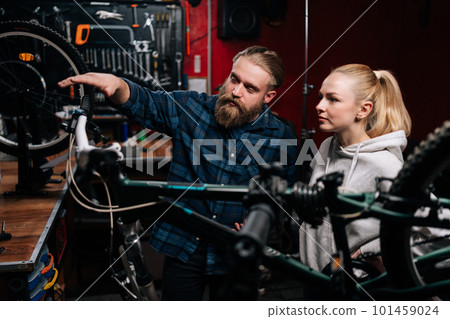 Bearded cycling repairman communicating with blonde female client, talking about problem of bicycle detected during diagnostics in repair shop with dark interior. Concept of bike maintenance. 101459024