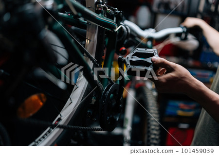 Close-up cropped shot of unrecognizable repairman holding the pedal of bicycle, insisting speed switches on mountain bike handlebar in repair shop with dark interior. Concept of maintenance of bike. 101459039