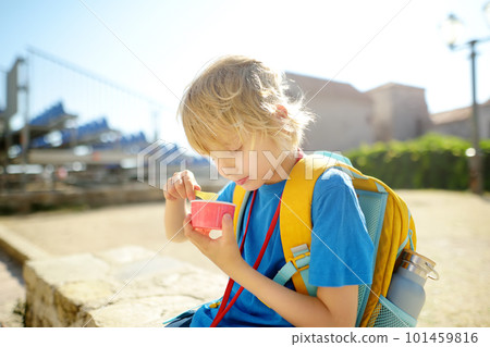 Happy little boy eating tasty ice cream in paper cup outdoors. School child have ice-cream snack on the go after school on sunny day. Kids love gelato 101459816
