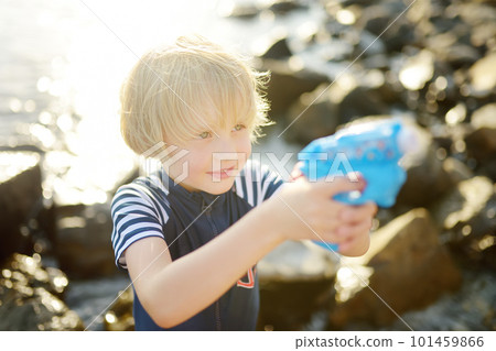 Cute little boy playing with water pistol on the seashore on a hot day. The child having fun with water gun outdoors during the summer holidays. 101459866