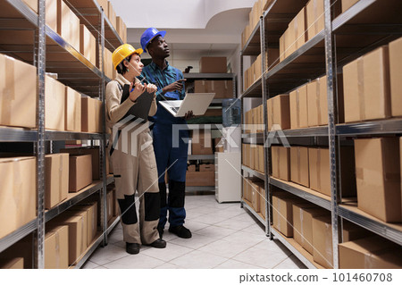 Shipping company workers managing goods stock control in warehouse, holding laptop and looking at parcels on shelf. Storehouse operators wearing protective helmets and overalls discussing dispatching 101460708