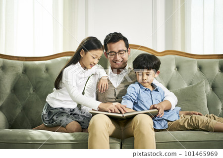 young asian father and two children sitting on couch reading book together in family living room at home young asian father and two children sitting on couch reading book together in family living room at home 101465969