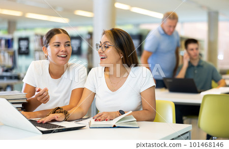 Two young women laughing and speaking loudly while using laptop in the library 101468146